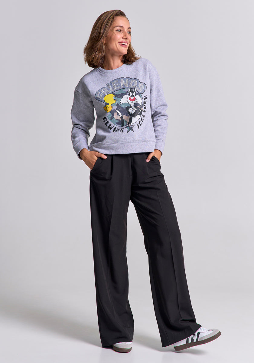 A woman with shoulder-length brown hair smiles with her hands in the pockets of her loose black pants, wearing a gray Frogbox by Princess Sylvester &amp; Tweety sweatshirt and gray sneakers, standing against a plain light gray background.