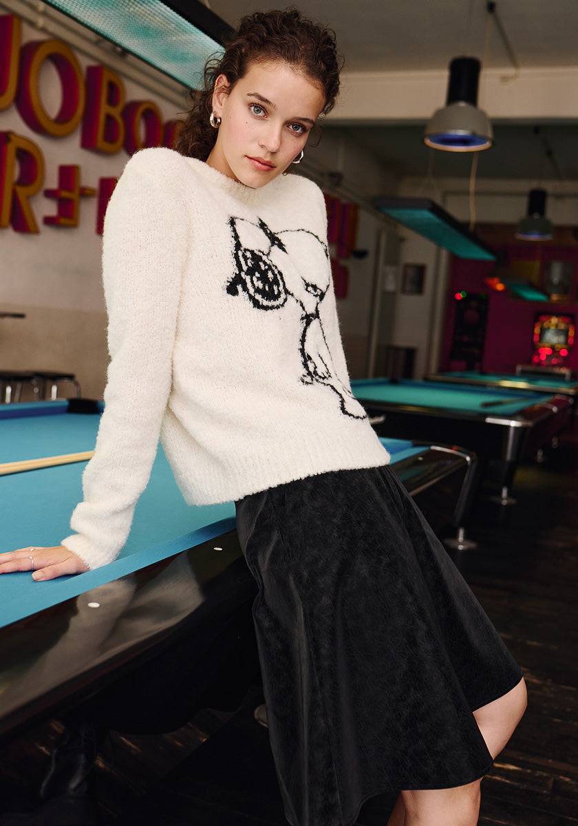 A young woman with curly hair leans against a blue pool table in a retro room, wearing a white Frogbox by Princess Grobstrickpullover with a black abstract face design and a black skirt. Neon signs and hanging lights glow behind her.