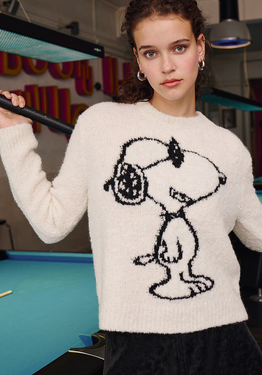 A young woman with curly hair, wearing hoop earrings and a cozy Frogbox by Princess Grobstrickpullover with a black Snoopy-with-headphones outline, stands by an indoor blue pool table holding a cue. Neon letters blur in the background.