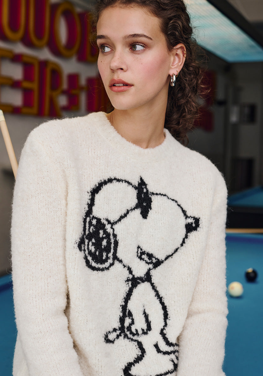 A young woman with curly brown hair wears a white Frogbox by Princess Snoopy Grobstrickpullover and stands by a blue pool table, looking left, with colorful wall signs and pool balls in the blurred background.