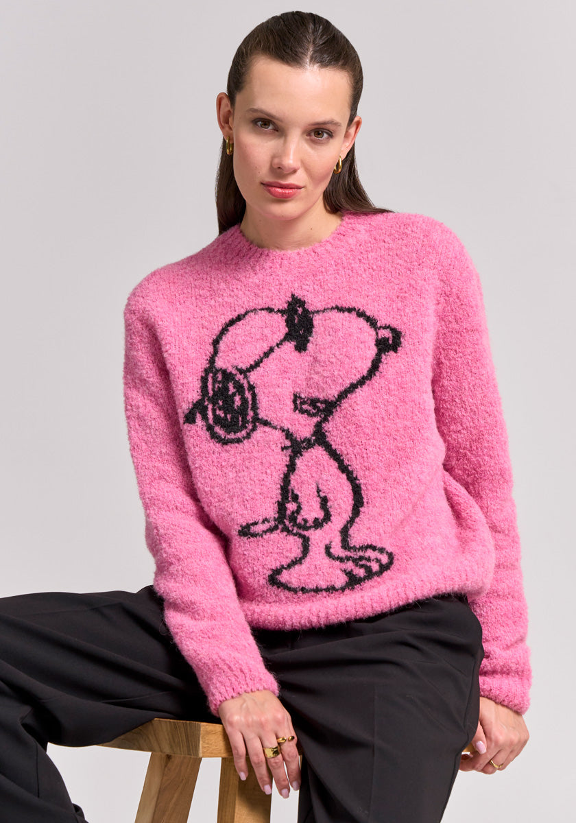 A woman with straight brown hair sits on a wooden stool, wearing a Frogbox by Princess Snoopy Grobstrickpullover aus Alpaka-Wolle-Mix and black pants. Gold hoop earrings and rings finish her confident look against a plain background.