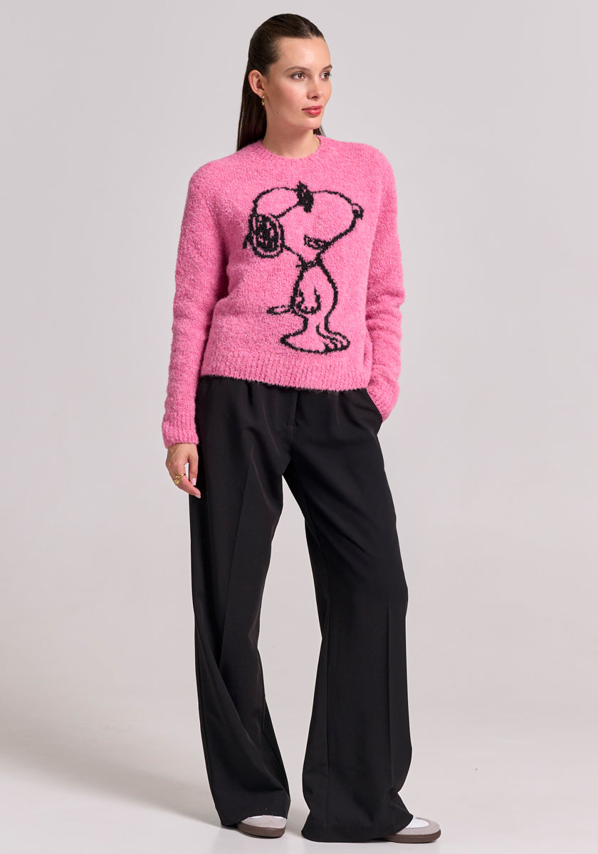 A woman with tied-back brown hair stands against a light gray background, wearing loose black trousers and a bright pink Frogbox by Princess Grobstrickpullover aus Alpaka-Wolle-Mix with a large black outline drawing of Snoopy.