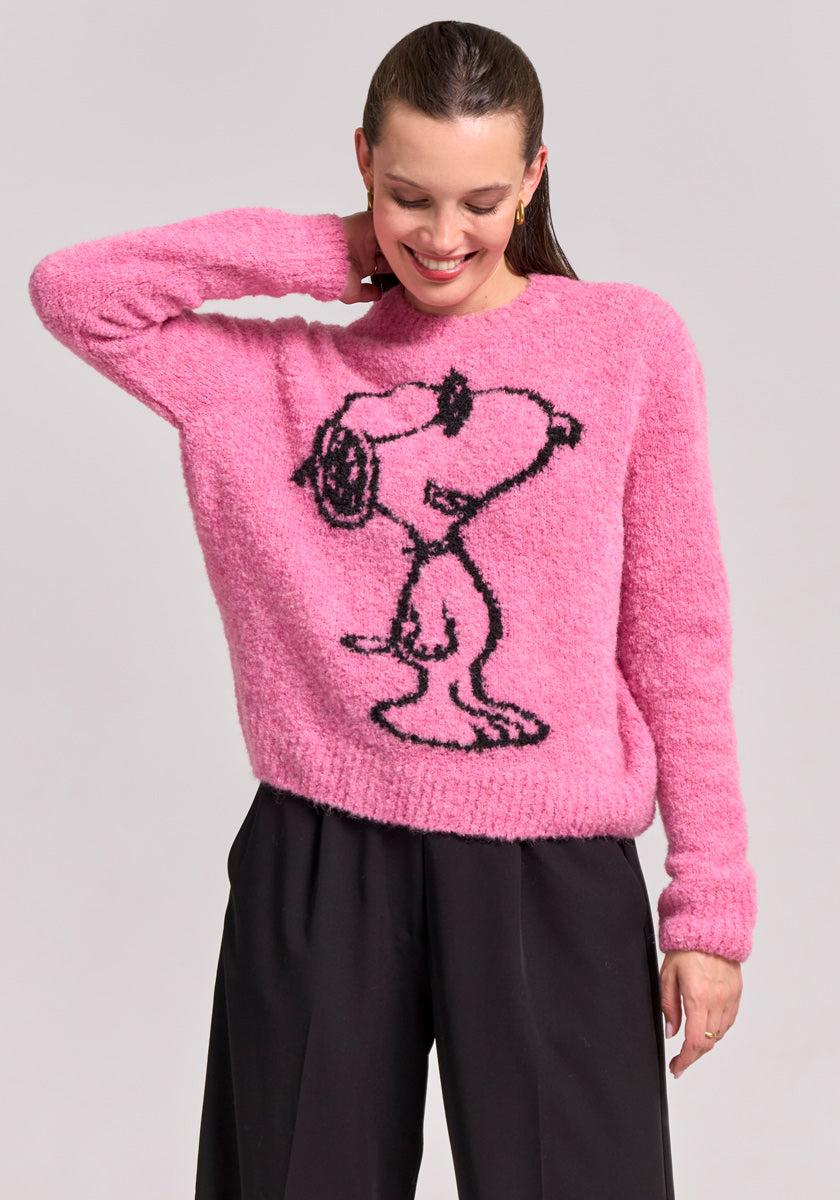 A smiling woman with slicked-back brown hair poses in a fuzzy pink Frogbox by Princess Grobstrickpullover featuring a large black outline of Snoopy from Peanuts and loose black pants, set against a plain light gray background.