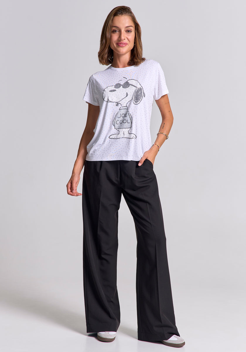 A woman with shoulder-length brown hair smiles against a plain background. She wears a Frogbox by Princess Snoopy "Joe Cool" T-shirt, loose black pants, and gray sneakers, with one hand in her pocket for a relaxed look.