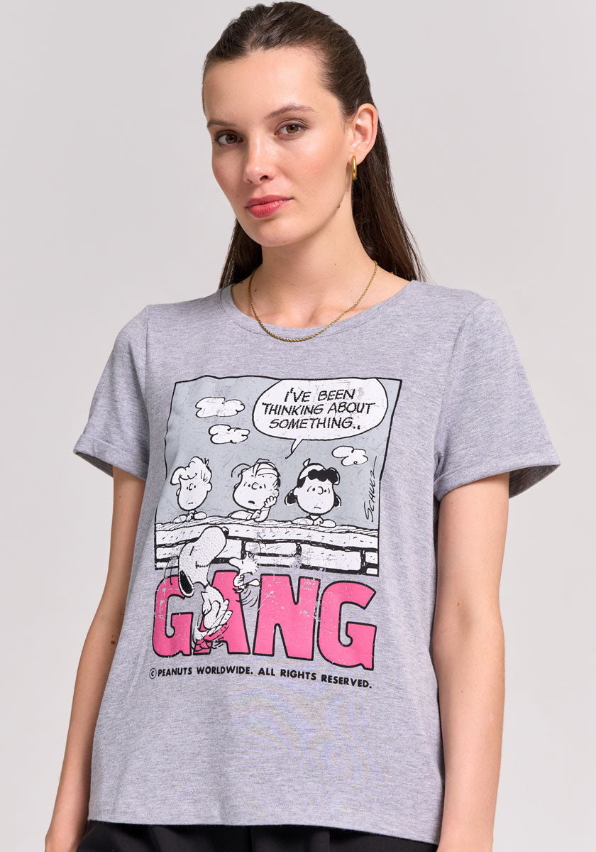 A woman with straight brown hair wears small gold hoop earrings, a gold necklace, and a Frogbox by Princess Peanuts-Print T-Shirt mit Stretch featuring Charlie Brown and GANG in bold pink letters, standing against a plain light background.
