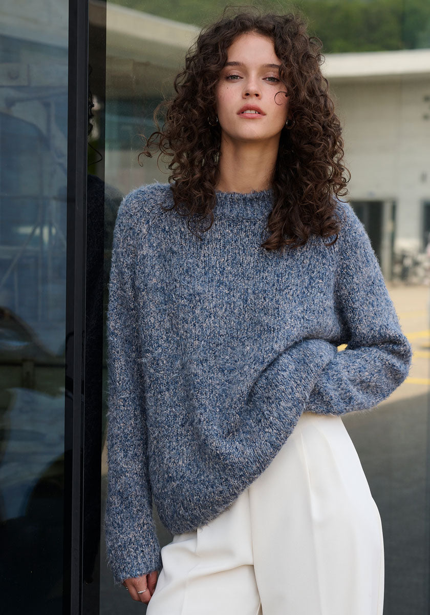Eine Frau mit lockigen braunen Haaren trägt einen blaugrauen Princess Goes Hollywood Strickpullover Seide mit Rundhalsausschnitt und eine hochtaillierte weiße Hose und steht im Freien vor einer Glaswand mit einem modernen Gebäude im Hintergrund.