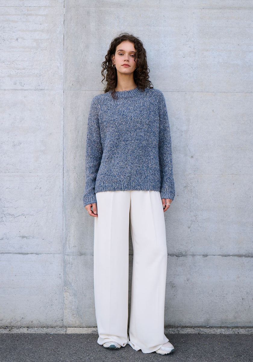 Eine Frau mit lockigem braunem Haar steht vor einer schlichten Betonwand und trägt einen blaugrauen Princess Goes Hollywood-Seidenpullover mit Rundhalsausschnitt und eine weite, hochgeschlossene weiße Hose. Ihre Arme liegen entspannt an den Seiten, und sie hat einen neutralen Gesichtsausdruck.