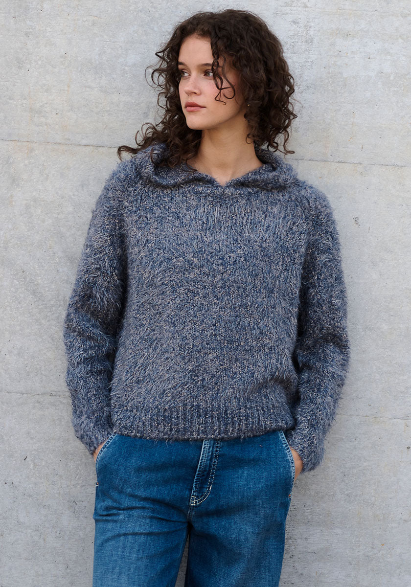 Eine Frau mit lockigem braunem Haar trägt eine graue Strickhoodie Seide von Princess Goes Hollywood und blaue Jeans. Sie steht an einer hellgrauen Betonwand, die Hände in den Taschen, und blickt mit neutraler Miene zur Seite.