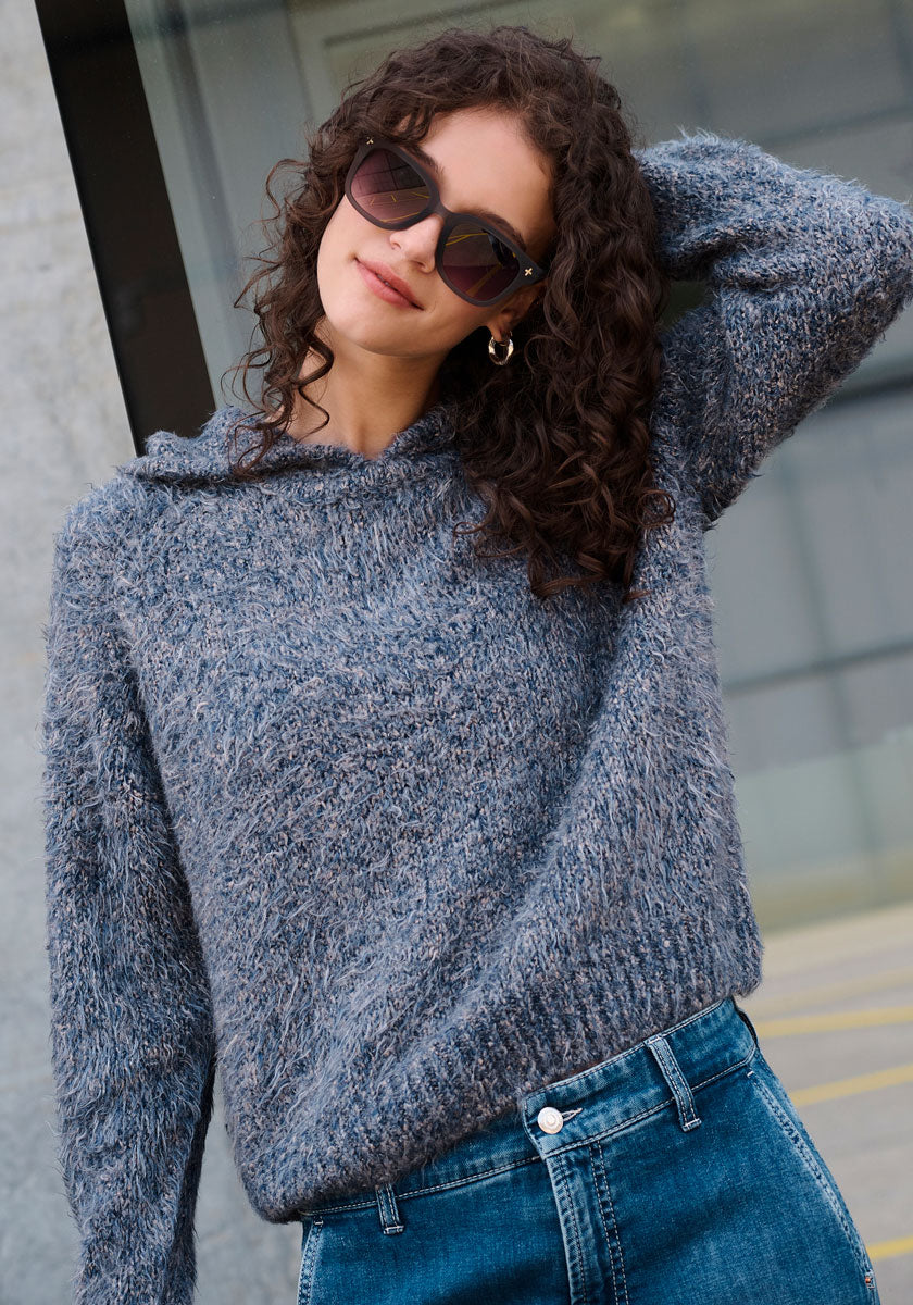 Eine Frau mit lockigem braunem Haar und dunkler Sonnenbrille trägt eine Princess Goes Hollywood Strickhoodie Seide mit Raglanärmeln und Kapuze sowie eine hoch sitzende Bluejeans. Mit einem sanften Lächeln neigt sie den Kopf und posiert vor einem Glasfenster und einer Steinwand.