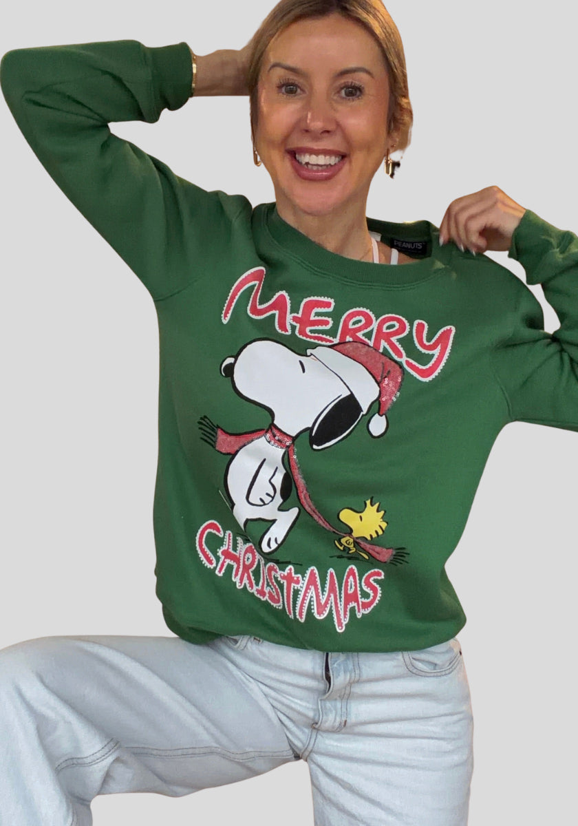 Eine lächelnde Frau posiert in einem Princess Goes Hollywood Snoopy Pullover, auf dem Snoopy mit Weihnachtsmannmütze, Woodstock und "Frohe Weihnachten" in festlichem Rot und Weiß abgebildet ist. Dazu trägt sie eine hellblaue Jeans und goldene Reifen vor einem grauen Hintergrund.