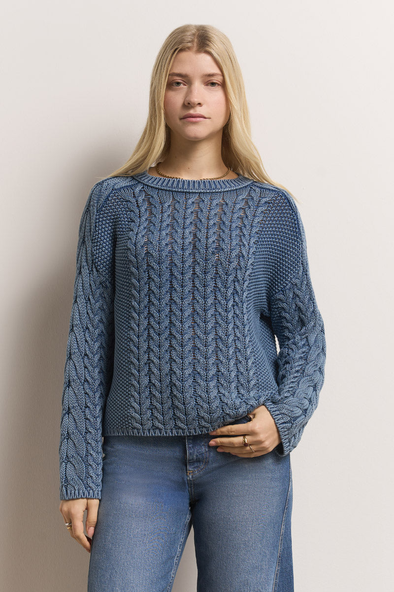 Eine junge Frau mit langen blonden Haaren trägt einen blauen Baumwollpullover mit langen Ärmeln aus Zopfstrick von Princess Goes Hollywood. Sie steht vor einem cremefarbenen Hintergrund und blickt neutral in die Kamera, gepaart mit einer blauen Jeans.