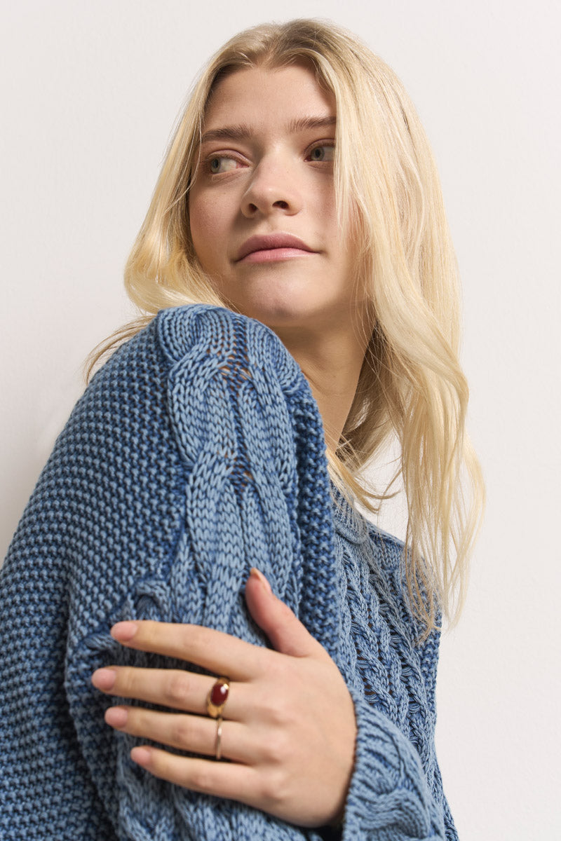 Eine Frau mit langen, gewellten blonden Haaren trägt einen blauen Zopfstrick Pullover aus Baumwolle von Princess Goes Hollywood. Sie blickt ruhig zur Seite, ihre Hand mit einem goldenen Ring und rotem Stein ruht sanft auf ihrem Pullover vor weißem Hintergrund.