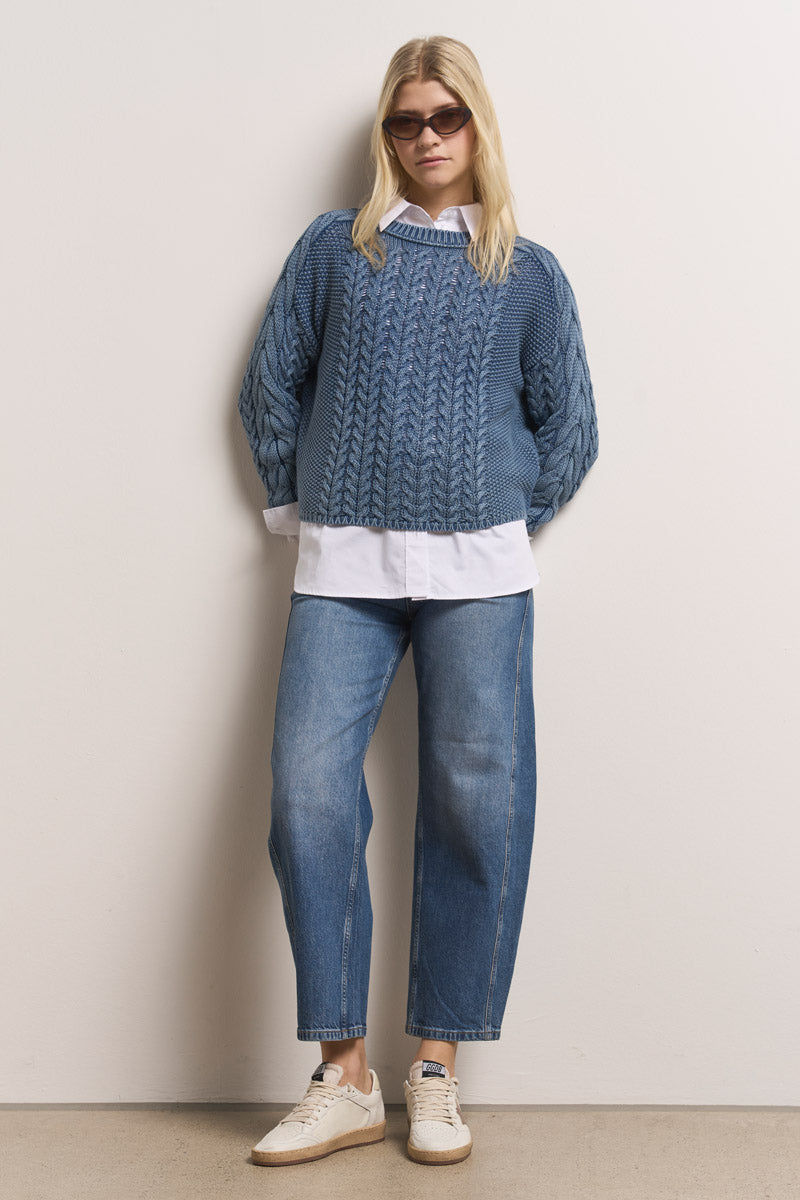 Eine Person mit langen blonden Haaren trägt eine Sonnenbrille, einen Princess Goes Hollywood hellblauen Zopfstrick Pullover aus Baumwolle über einem weißen Button-up, lässige Blue Jeans im Stone-Wash-Effekt und weiße Sneakers. Sie stehen entspannt vor einer beigen Wand.