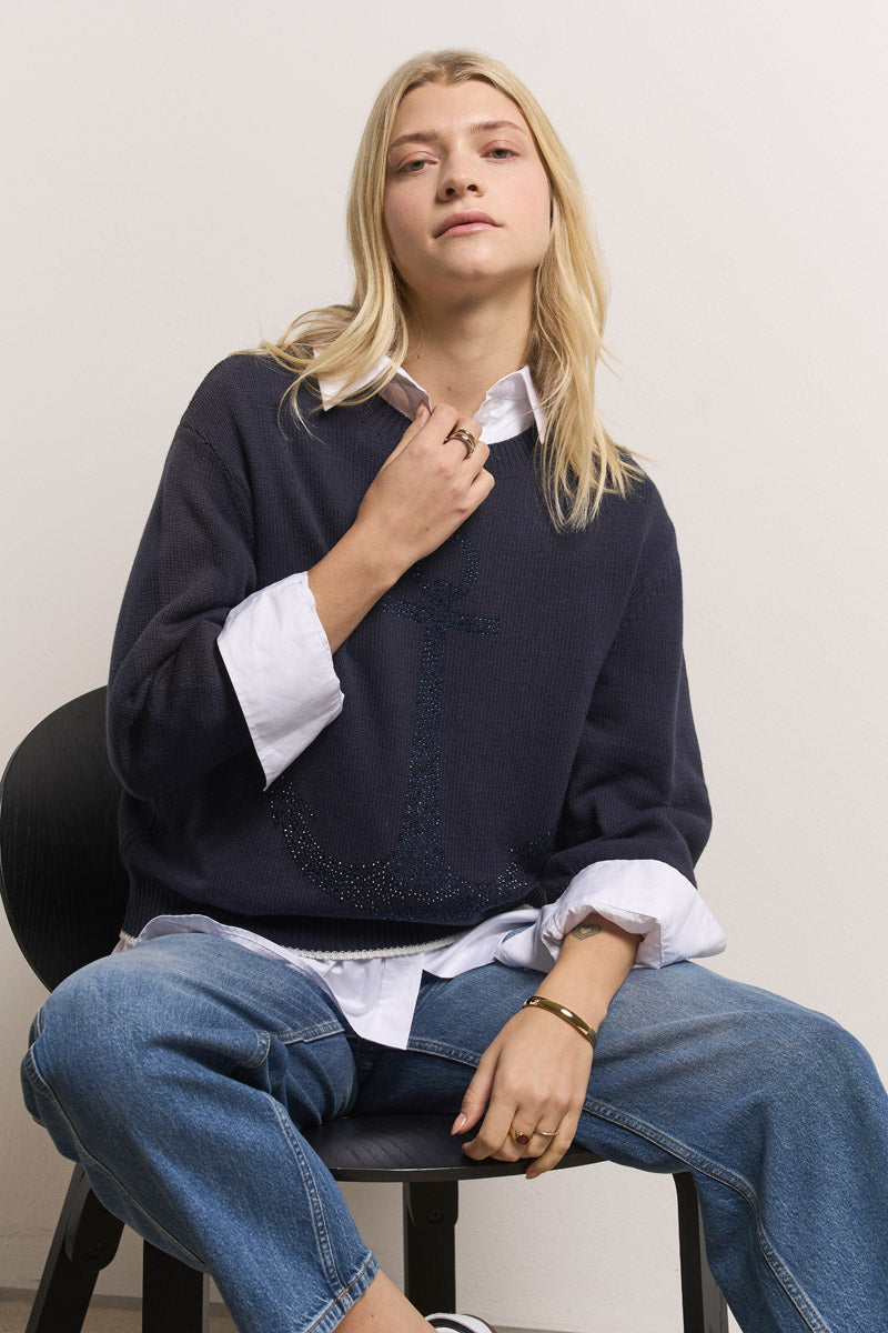 Eine Frau mit langen blonden Haaren sitzt auf einem schwarzen Stuhl, trägt einen marineblauen Princess Goes Hollywood Baumwolle-Mix Pullover mit Strassverzierung über einem weißen Hemd mit Kragen und blauen Jeans und blickt selbstbewusst und in entspannter Haltung in die Kamera.