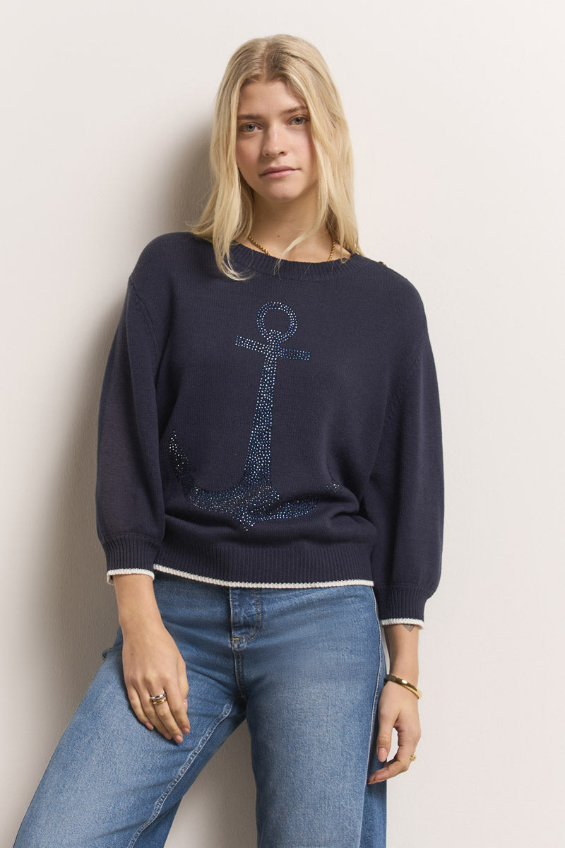 Eine Frau mit langen blonden Haaren trägt einen marineblauen Princess Goes Hollywood Baumwolle-Mix Pullover mit Anker-Motiv aus Strassverzierung, gepaart mit hellblauen Jeans und Ringen, steht vor einer cremefarbenen Wand und schaut ruhig in die Kamera.