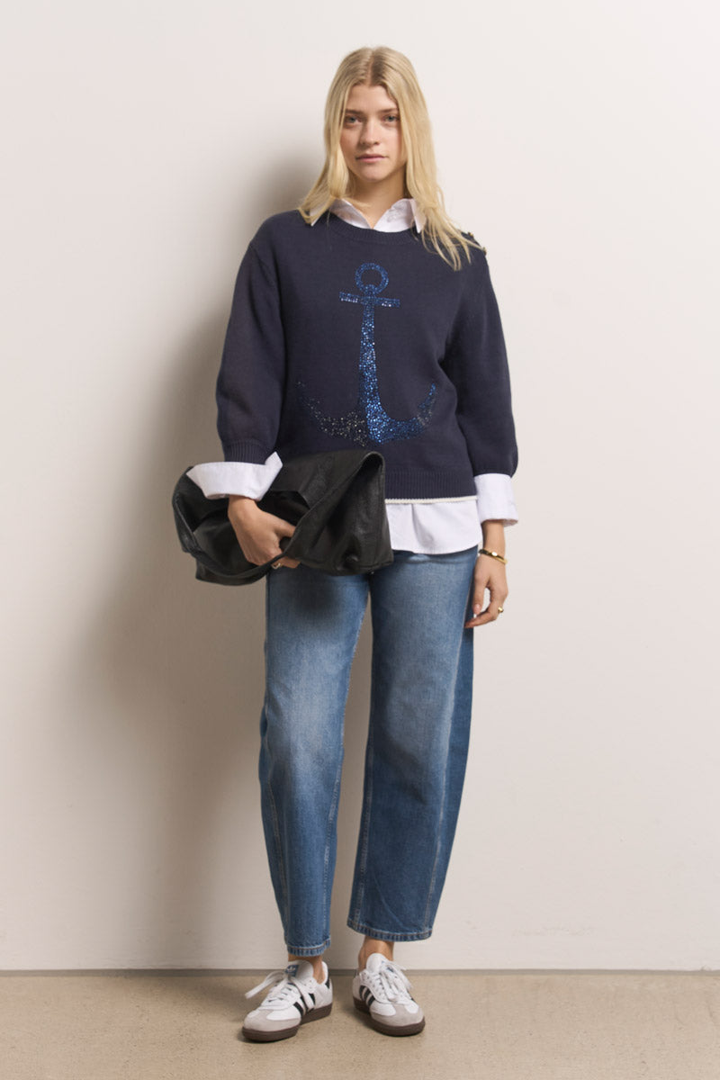 Eine blonde Frau trägt einen marineblauen Baumwolle-Mix-Pullover mit einem großen blauen Ankermotiv von Princess Goes Hollywood über einem weißen Hemd, kombiniert mit blauen Jeans, weißen Turnschuhen und einer schwarzen Clutch, während sie vor einer hellen Wand auf einem beigen Boden steht.