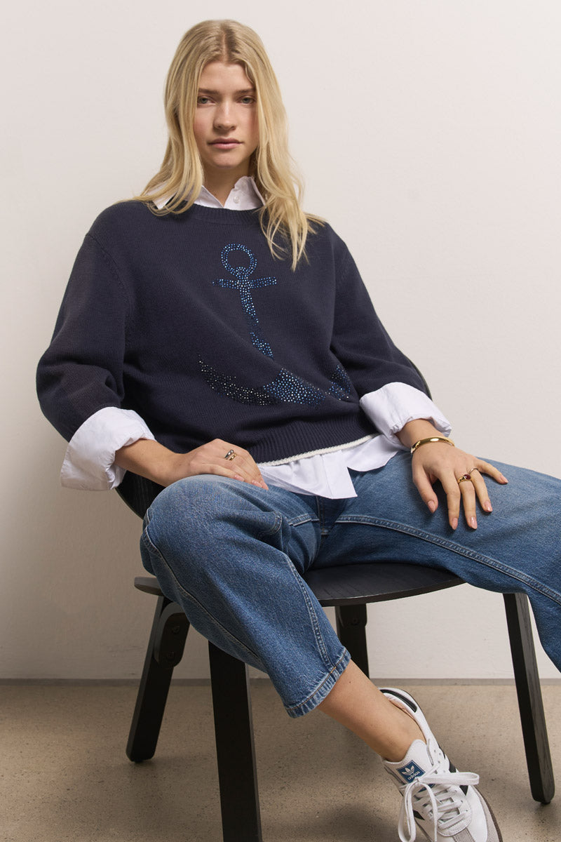 Eine Frau mit langen blonden Haaren sitzt auf einem schwarzen Stuhl und trägt einen marineblauen Princess Goes Hollywood Baumwolle-Mix Pullover mit Strassverzierung und Anker-Motiv über einem weißen Shirt, blaue gekrempelte Jeans und weiße Sneakers. Sie sieht entspannt und selbstbewusst aus.