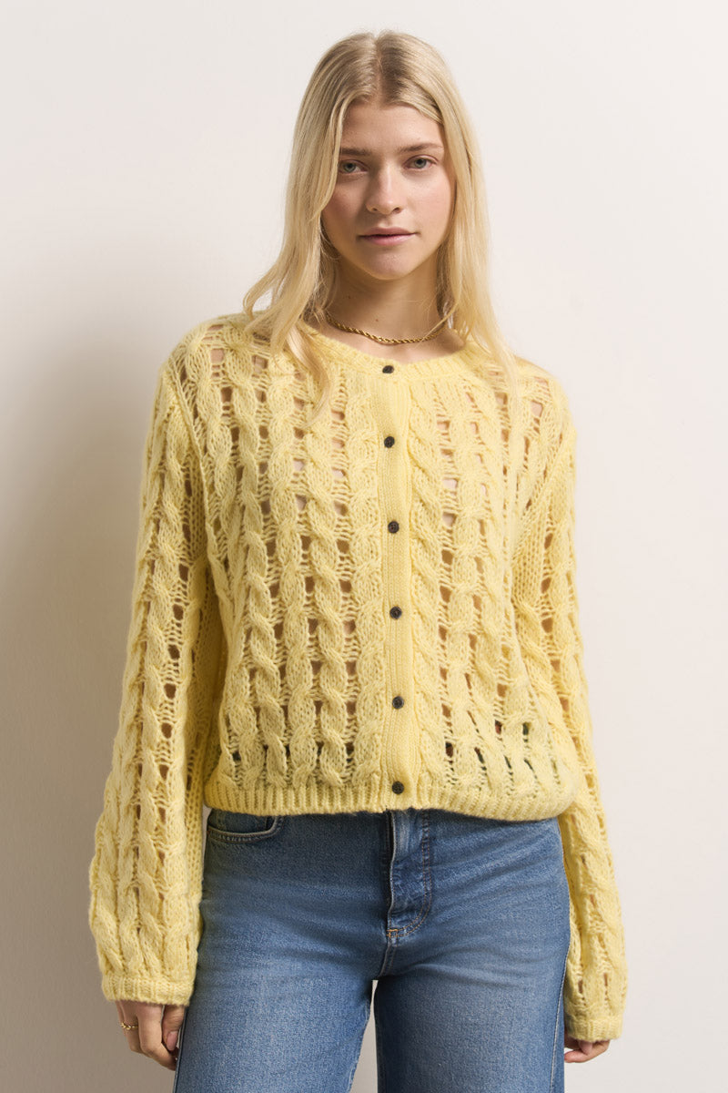 Eine junge Frau mit langen blonden Haaren trägt eine hellgelbe Merino-Alpaka-Mix Lochstrickjacke von Princess Goes Hollywood über einem beigen Oberteil, blauen Jeans und einer dezenten Halskette. Sie steht vor einem weißen Hintergrund und schaut ruhig in die Kamera.