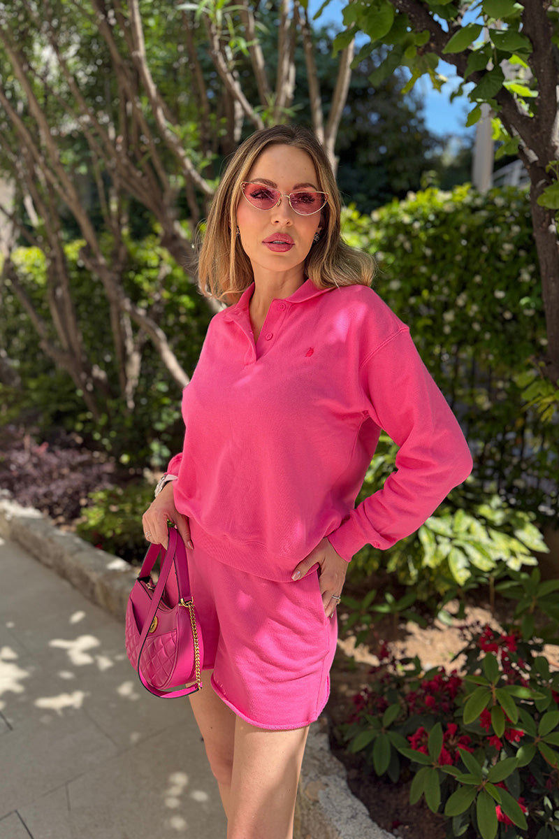 Eine Frau steht draußen inmitten von Grün und trägt ein leuchtend pinkfarbenes „Princess Goes Hollywood“-Sweatshirt aus Baumwolle sowie dazu passende Shorts. Dazu trägt sie eine pinkfarbene Handtasche und eine getönte Sonnenbrille; ihr farbenfroher Look wird durch das durch die Bäume fallende Sonnenlicht besonders hervorgehoben.