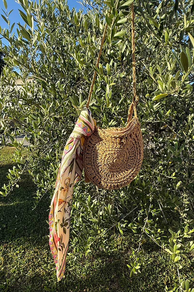 Eine runde Umhängetasche aus Stroh von Princess Goes Hollywood mit gewebter Struktur hängt an einem Baumzweig, der mit einem farbenfrohen Blumenschal am Henkel gestylt ist. Das schicke Accessoire hebt sich von üppig grünen Blättern und einem blauen Himmelshintergrund ab.