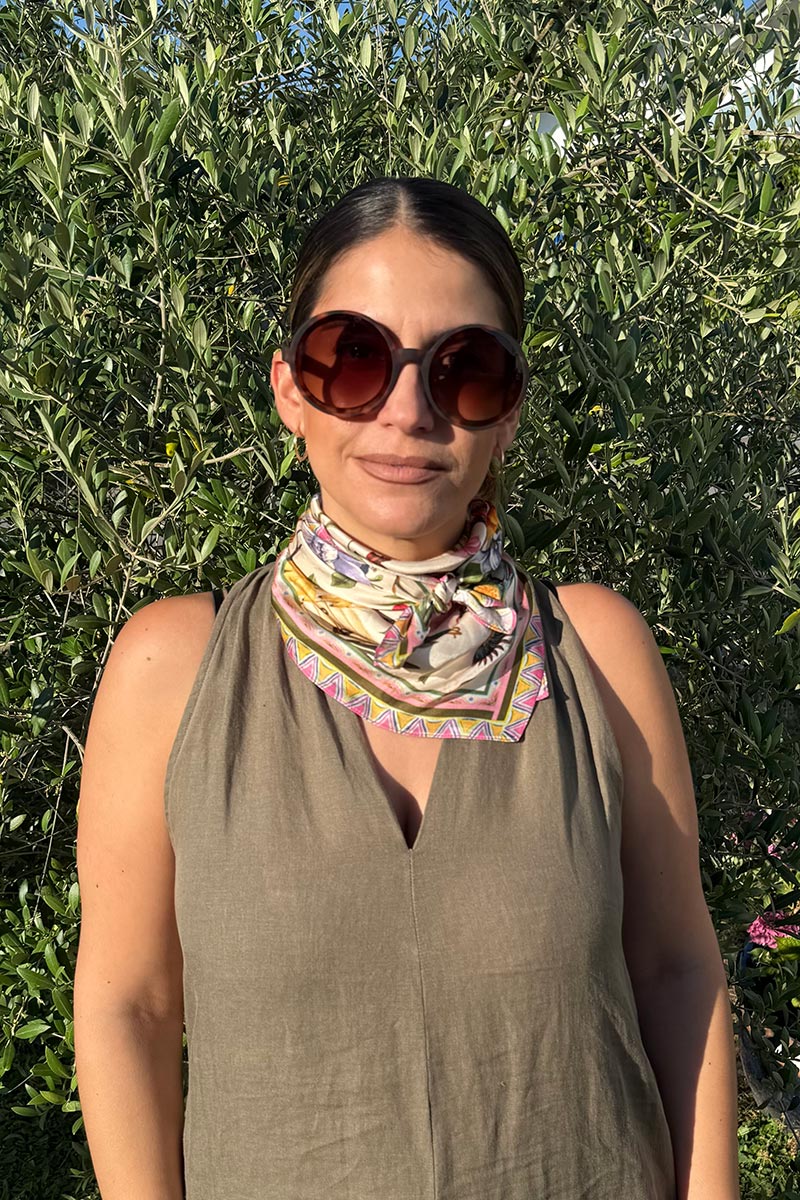 Eine Frau mit dunklem, zurückgestecktem Haar trägt eine große runde Sonnenbrille, ein ärmelloses olivgrünes Top von Princess Goes Hollywood und ein bunt gemustertes Tuch als Styling-Highlight. Sie steht vor üppig grünem Laub an einem sonnigen Tag.