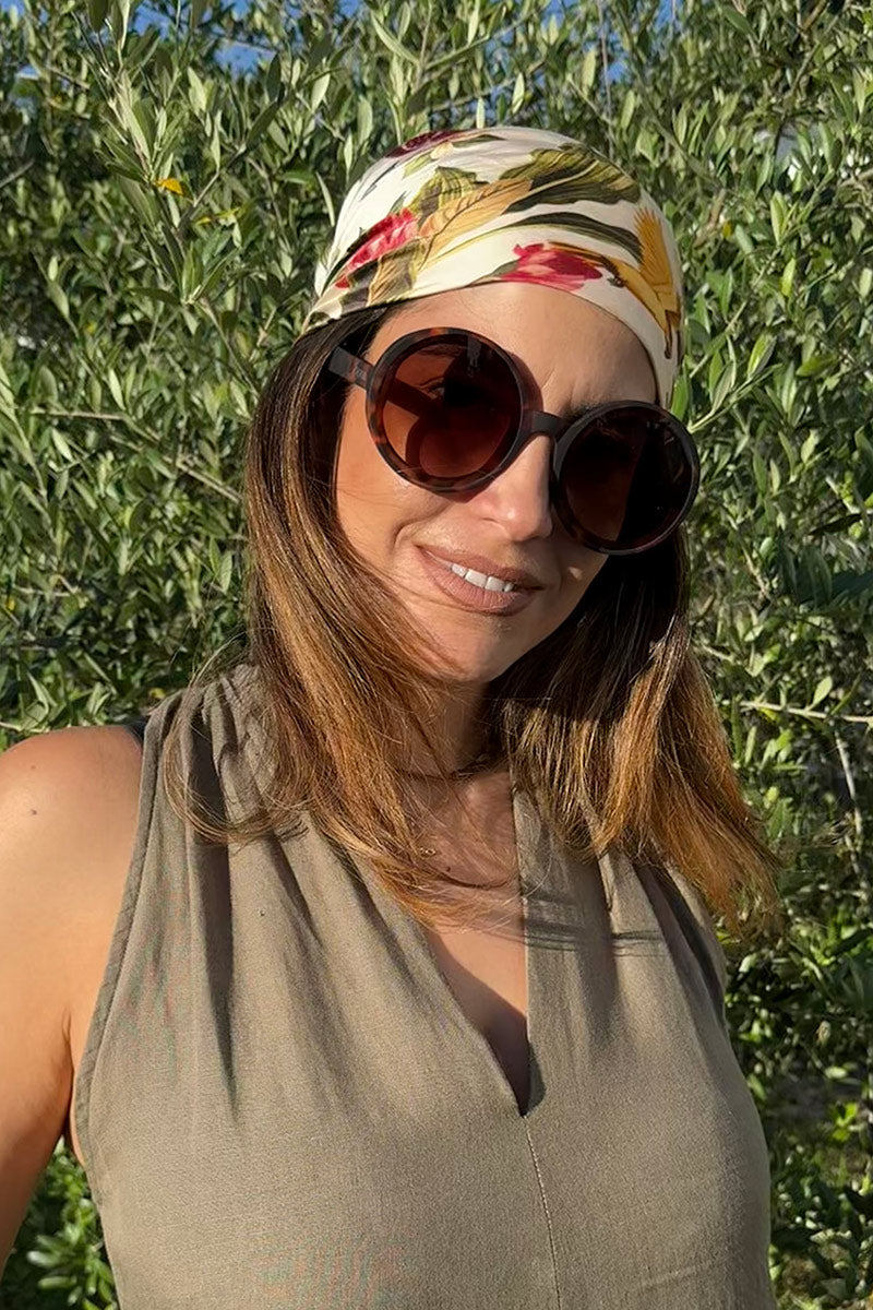 Eine Frau lächelt im Freien, trägt eine große runde Sonnenbrille und ein geblümtes Princess Goes Hollywood-Tuch als Kopftuch. Das Accessoire passt zu ihren braunen Haaren und ihrem beigefarbenen ärmellosen Oberteil, das im Sonnenlicht mit grünen Blätterzweigen im Hintergrund hervorgehoben wird.