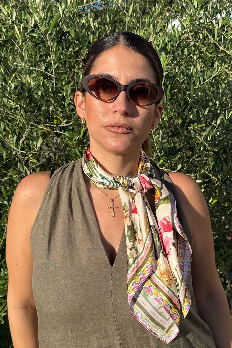 Eine Frau steht im Freien vor belaubten Büschen und trägt die ovale braune Sonnenbrille von Princess Goes Hollywood, ein ärmelloses olivgrünes Top und ein buntes Blumentuch als schickes Accessoire, gepaart mit goldenen Reifen und einer zarten Halskette.