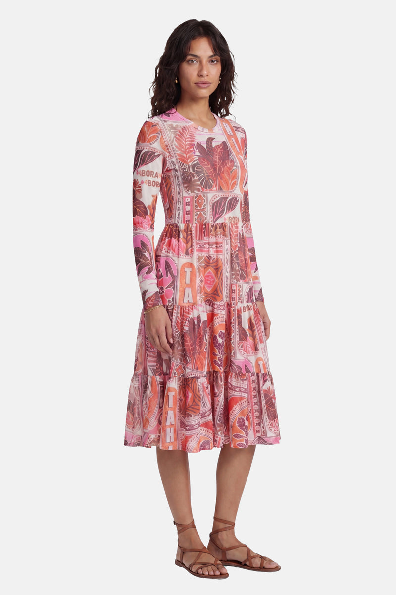 Eine Frau mit welligem, dunklem Haar trägt ein langärmeliges Netzkleid der Marke „Princess Goes Hollywood“ mit einem auffälligen rosa Tahiti-Print – Blätter, abstrakte Formen und der Schriftzug „TAHITI“ –, kombiniert mit braunen Riemchensandalen ohne Absatz vor einem schlichten weißen Hintergrund.