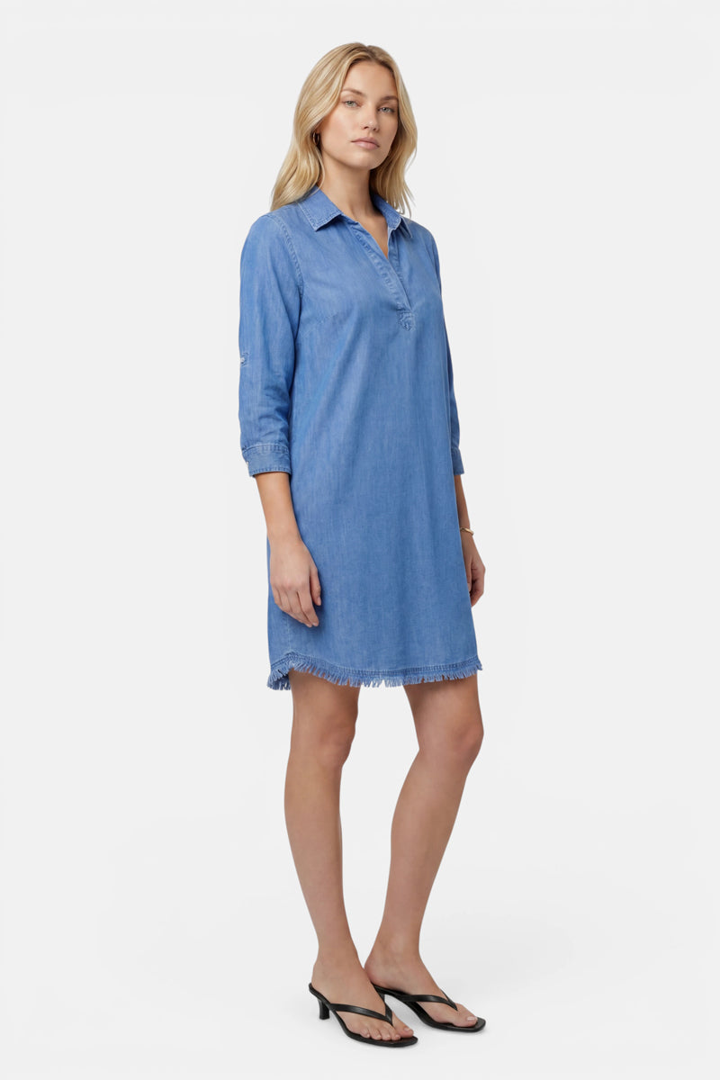 Eine Frau mit blonden Haaren und ausdruckslosem Gesicht trägt ein locker sitzendes, hellblaues Lyocell-Blusenkleid von Princess Goes Hollywood mit Hemdkragen und ausgefranstem Saum. Dazu trägt sie schwarze Sandalen vor einem schlichten weißen Hintergrund.