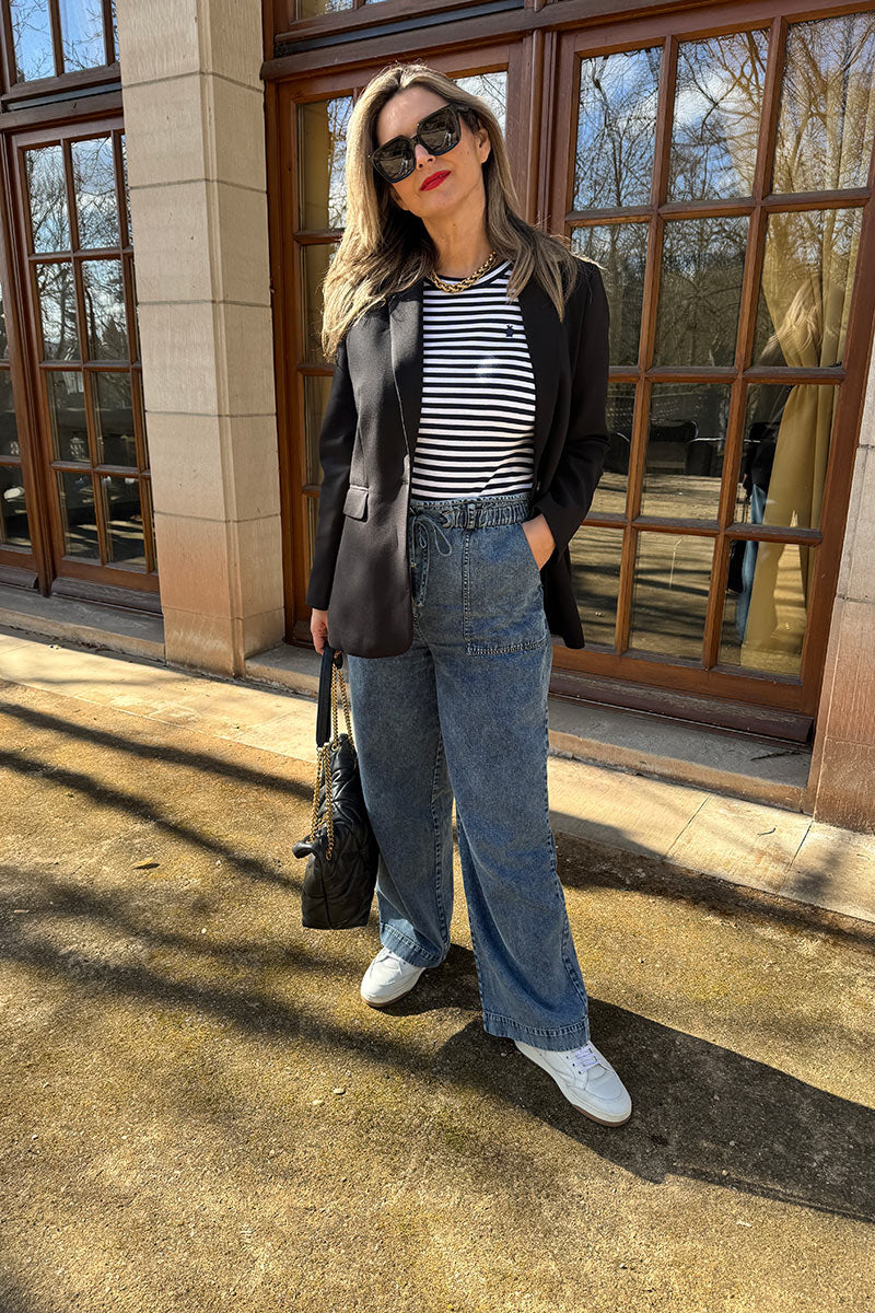 Eine Frau steht im Freien im Sonnenlicht und trägt eine schwarze Sonnenbrille, einen schwarzen Blazer, ein schwarz-weiß gestreiftes Hemd und eine weite Jeanshose aus Baumwolldenim mit Paperbag-Bund. Sie hält eine schwarze Handtasche neben einem Gebäude mit großen Fenstern.