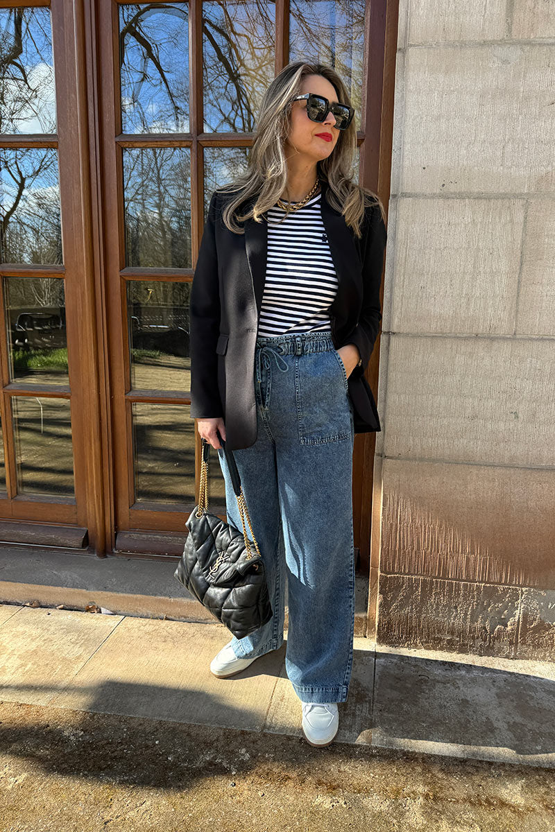 Eine Frau mit langem, gewelltem blondem Haar steht im Freien an einem großen Fenster. Sie trägt eine schwarze Sonnenbrille, einen schwarzen Blazer, ein gestreiftes Hemd, eine lockere Princess Goes Hollywood Baumwolldenim Jeanshose im Paperbag-Bund und weiße Turnschuhe, während sie eine schwarze gesteppte Tasche in der Hand hält.