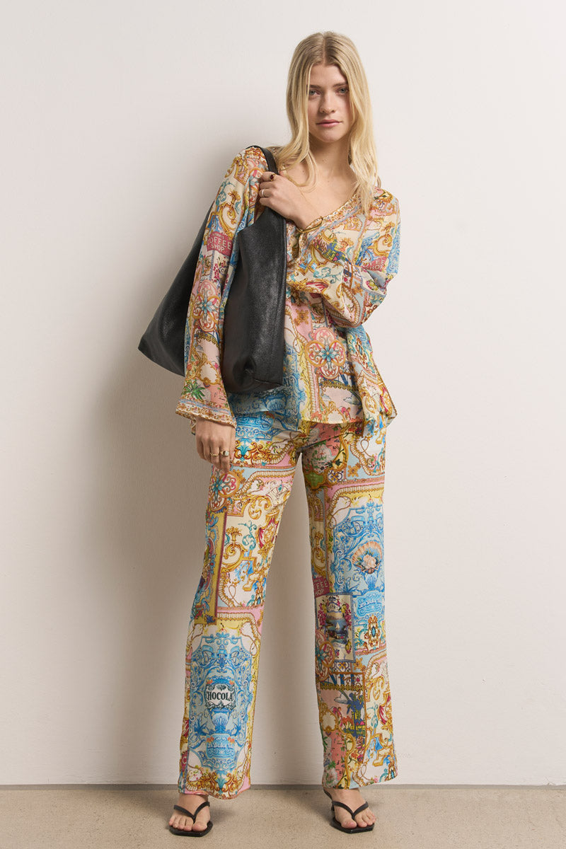 Eine Frau mit langen blonden Haaren steht vor einer weißen Wand. Sie trägt eine farbenfrohe Princess Goes Hollywood Dolce Vita-Print-Bluse und dazu passende blaue, rosa und goldene Seidenhosen mit Gummibund. Sie hält eine große schwarze Tragetasche in der Hand und trägt schwarze Sandalen.