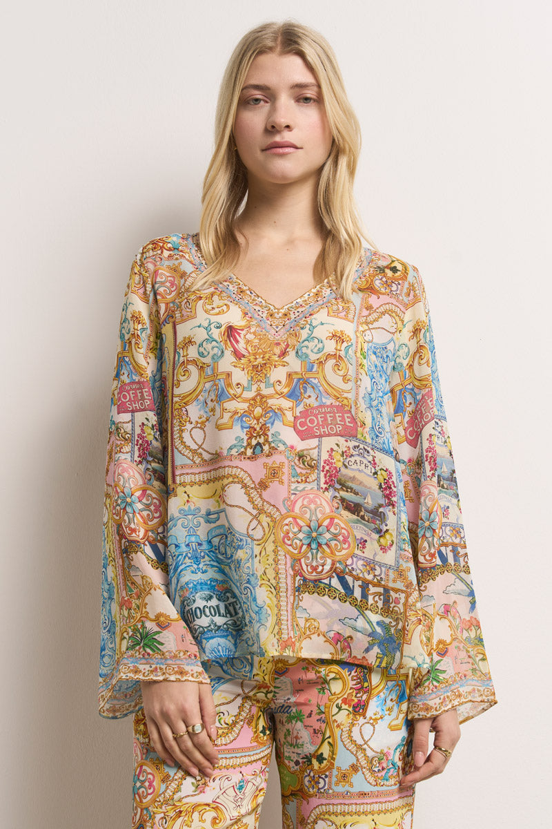 Eine Frau mit langen blonden Haaren trägt eine farbenfrohe Princess Goes Hollywood-Seidenbluse und eine dazu passende Hose, die beide mit Dolce Vita-Print-Motiven wie "Coffee Shop" und "Chocolate" in Pastellrosa, Blau, Gelb und Gold verziert sind. Sie steht vor einem schlichten hellen Hintergrund.