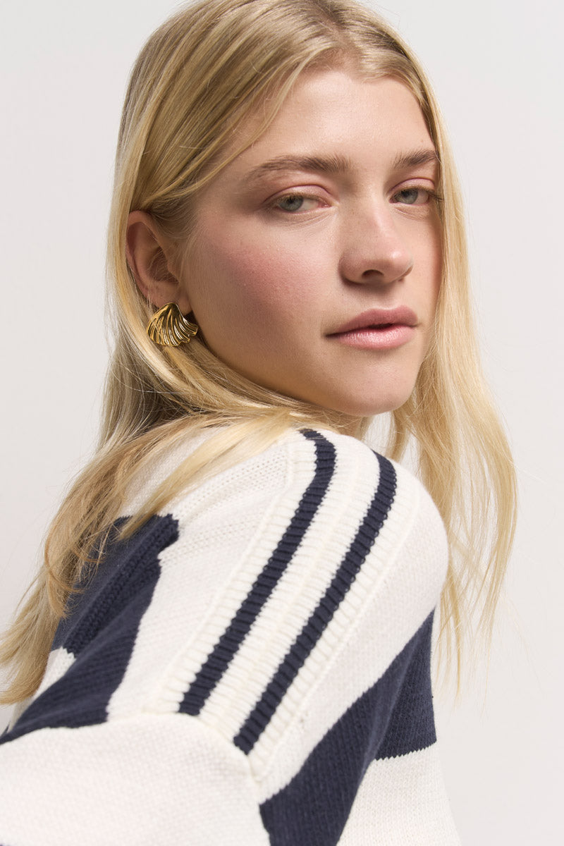 Eine junge Frau mit langen blonden Haaren trägt einen gestreiften Pullover aus Baumwollmix von Princess Goes Hollywood. Sie blickt über ihre Schulter in die Kamera, trägt goldene Ohrringe und minimales Make-up vor einem schlichten, hellen Hintergrund.