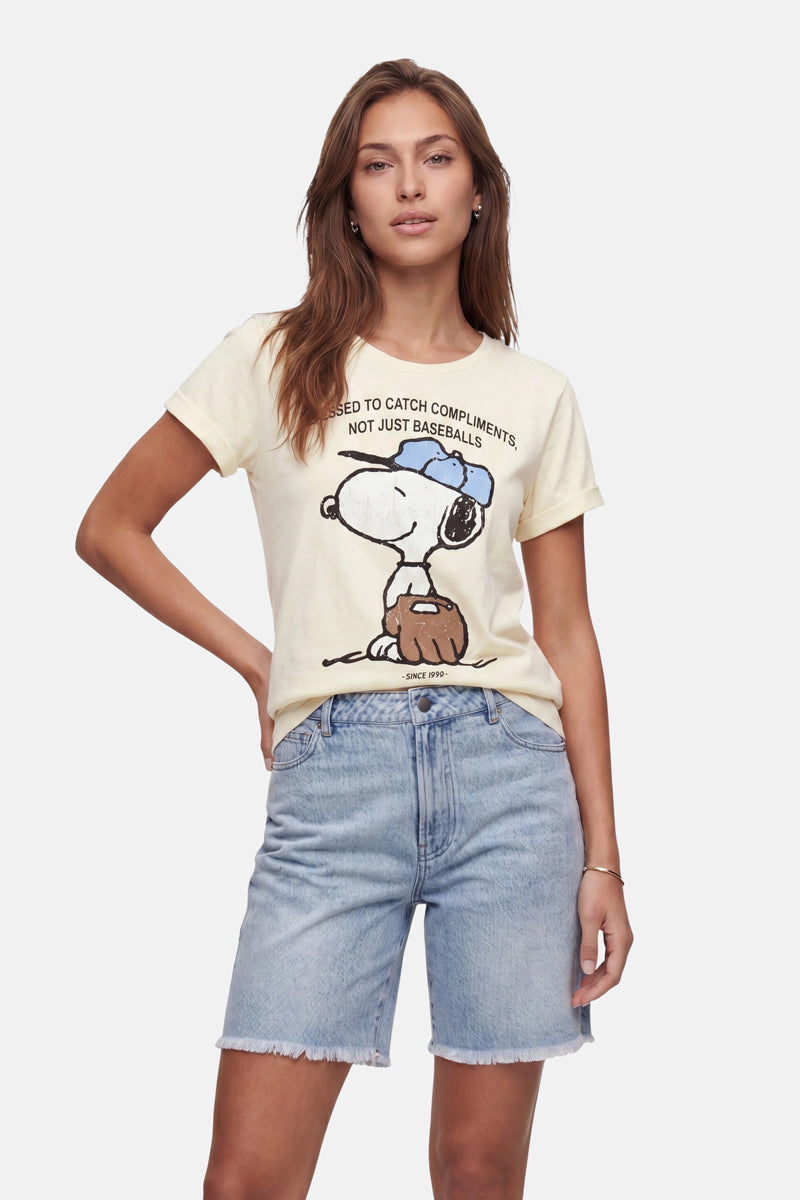 Vor einem schlichten Hintergrund steht eine junge Frau mit langen braunen Haaren. Sie trägt ein hellgelbes „Frogbox by Princess Limited Edition Snoopy-Print“ Baumwoll-T-Shirt und hellblaue Jeansshorts, eine Hand in die Hüfte gestützt, und strahlt eine entspannte Selbstsicherheit aus.