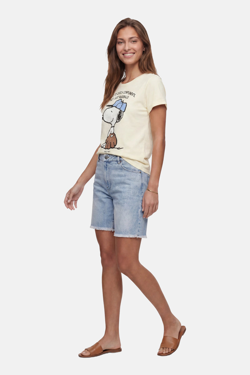 Eine Frau mit langen braunen Haaren lächelt. Sie trägt ein limitiertes „Frogbox by Princess Snoopy“-T-Shirt aus Baumwolle, hellblaue, knielange Jeansshorts und braune flache Sandalen. Sie steht vor einem schlichten weißen Hintergrund, die Hände in die Hüften gestemmt.