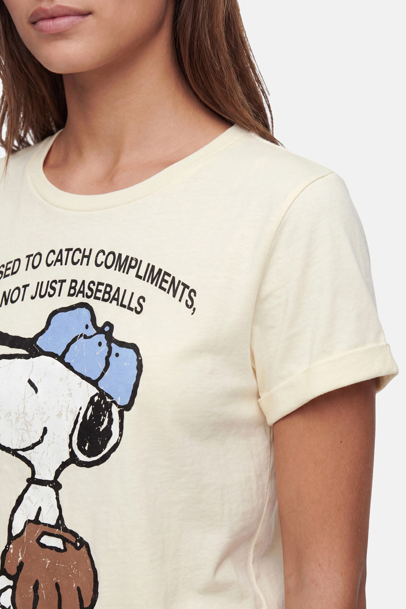 Eine Frau mit langen braunen Haaren trägt ein hellgelbes T-Shirt der „Frogbox by Princess Limited Edition“ mit Snoopy-Aufdruck, auf dem Snoopy mit blauem Hut und Handschuh zu sehen ist, zusammen mit dem Schriftzug „USED TO CATCH COMPLIMENTS, NOT JUST BASEBALLS“. Zu sehen sind nur ihr unterer Gesichtsbereich und ihr Oberkörper.