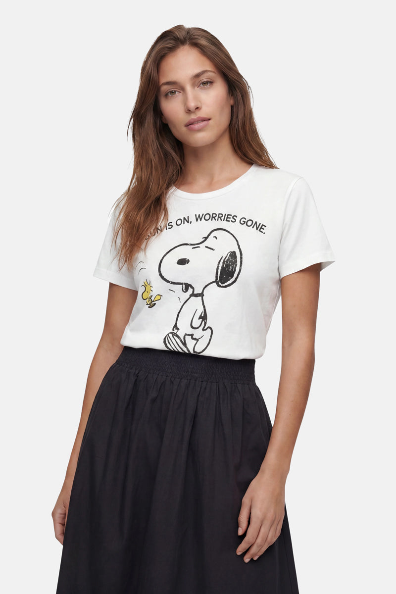 Eine Frau mit langen braunen Haaren trägt ein „Frogbox by Princess Limited Edition Snoopy“-T-Shirt aus Baumwolle mit Woodstock und dem Aufdruck „Always on, worries gone“. Dazu trägt sie einen schwarzen Rock mit hoher Taille vor einem hellen Hintergrund.