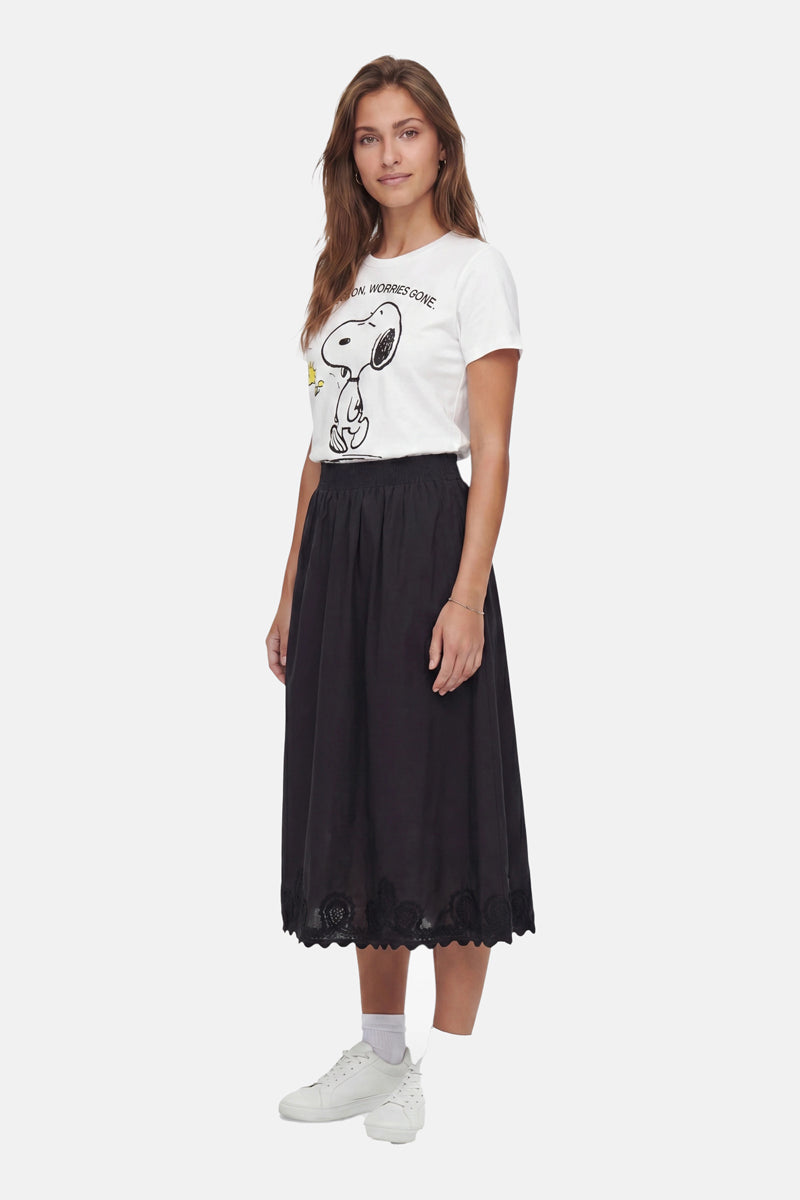 Eine Frau mit langen braunen Haaren lächelt sanft. Sie trägt ein weißes Snoopy-T-Shirt aus Baumwolle der „Frogbox by Princess Limited Edition“, einen schwarzen, mittellangen Rock mit Wellenrand, weiße Socken und Turnschuhe. Sie steht leicht nach links versetzt vor einem schlichten weißen Hintergrund.