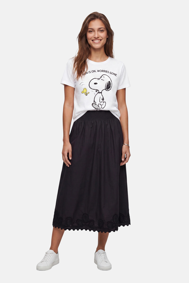 Eine Frau mit langen braunen Haaren lächelt in einem „Frogbox by Princess Limited Edition Snoopy“-T-Shirt aus Baumwolle, auf dem der Text „Mean is OK. Worries Gone.“ zu lesen ist. Dazu trägt sie einen schwarzen Midirock mit Spitzenbesatz und weiße Sneakers vor einem schlichten Hintergrund.