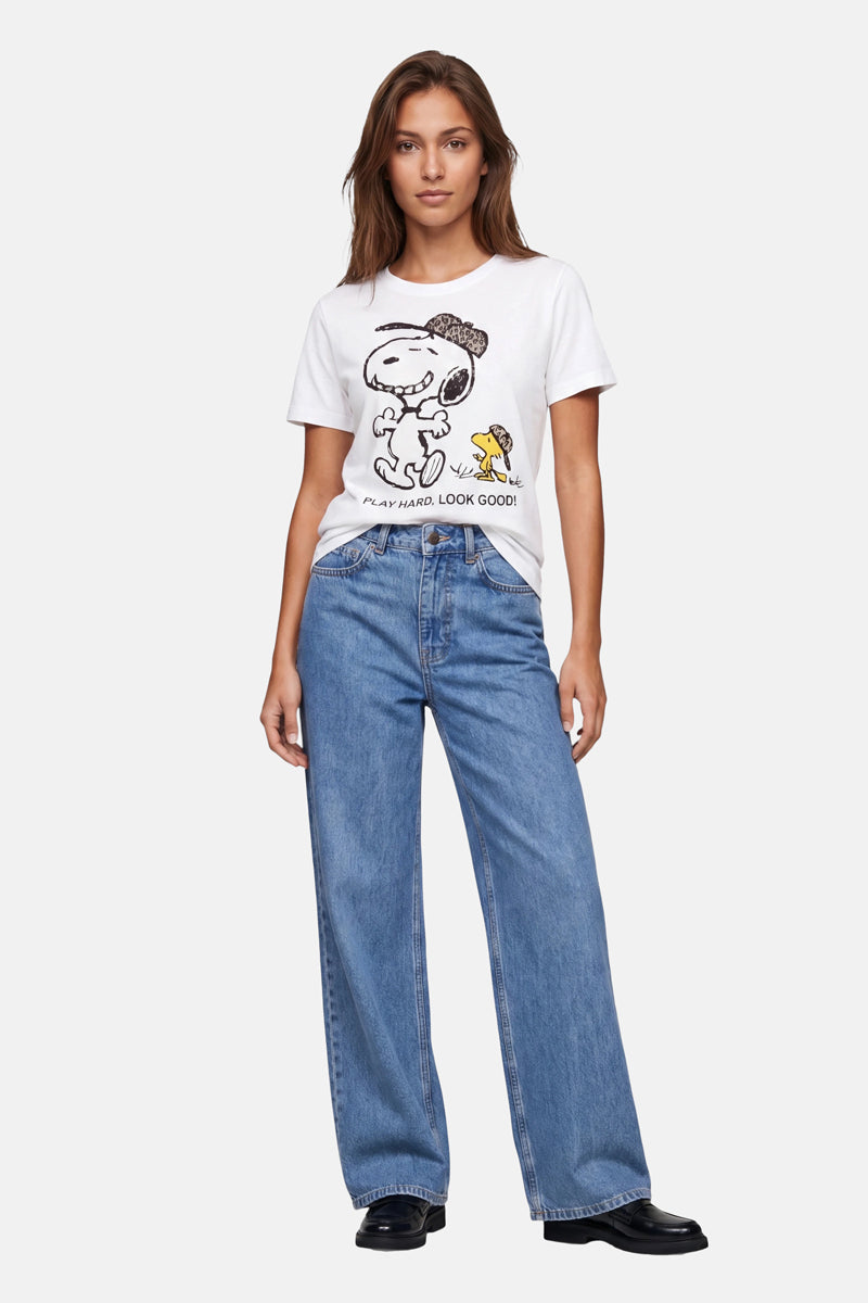 Eine Frau blickt nach vorne in einem „Frogbox by Princess Limited Edition Snoopy“-T-Shirt aus Baumwolle, auf dem Woodstock und der Schriftzug „Play Hard. Look Good!“ zu sehen sind. Dazu trägt sie eine lockere blaue Jeans und schwarze Schuhe. Ihr braunes Haar fällt ihr vor einem schlichten weißen Hintergrund ins Gesicht.