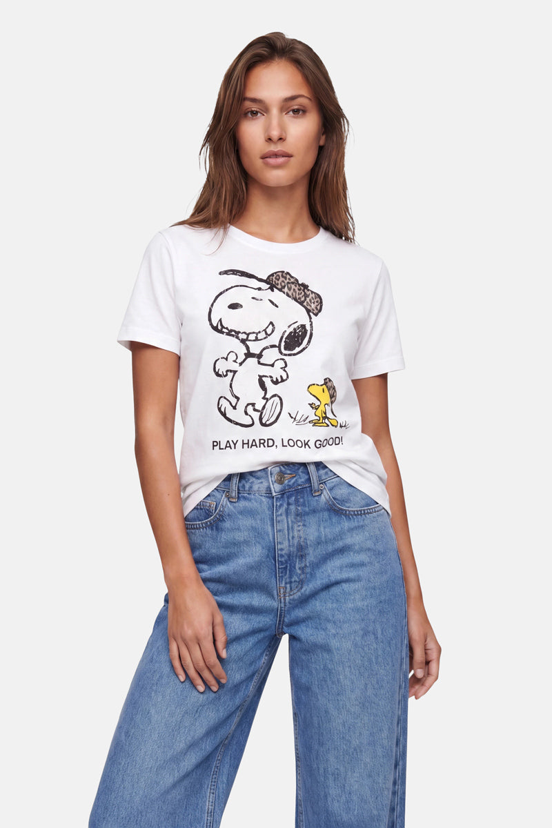 Eine Frau trägt ein weißes „Frogbox by Princess Snoopy“-T-Shirt aus weicher Baumwolle mit der Aufschrift „PLAY HARD, LOOK GOOD!“ und einem Woodstock-Aufdruck, dazu eine blaue Jeans. Sie steht vor einem einfarbigen Hintergrund, die linke Hand lässig in der Hosentasche.
