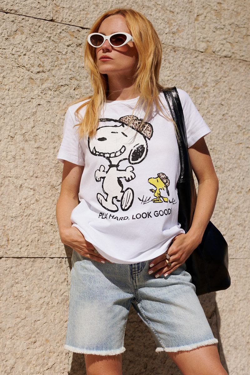 Eine Frau mit langen blonden Haaren trägt eine weiße Sonnenbrille im Cat-Eye-Stil, ein „Frogbox by Princess Limited Edition Snoopy“ T-Shirt aus Baumwolle mit der Aufschrift „PLAY HARD. LOOK GOOD!“, helle Jeansshorts und eine große schwarze Tasche; sie steht vor einer beigen Steinmauer.
