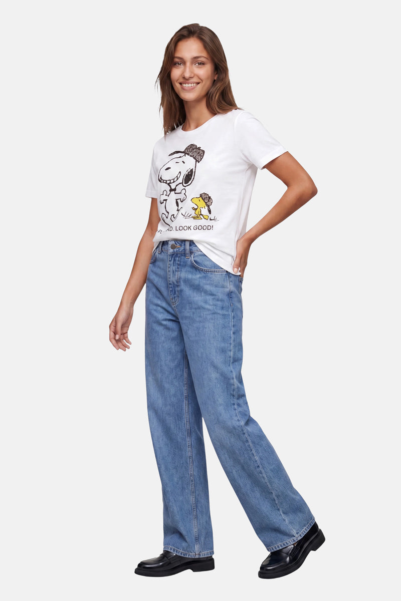 Eine Frau lächelt. Sie trägt ein „Frogbox by Princess Limited Edition Snoopy“-T-Shirt aus Baumwolle, eine blaue Jeans mit hoher Taille und weitem Bein sowie schwarze Schuhe. Vor einem schlichten hellgrauen Hintergrund posiert sie mit einer Hand in der Hüfte.