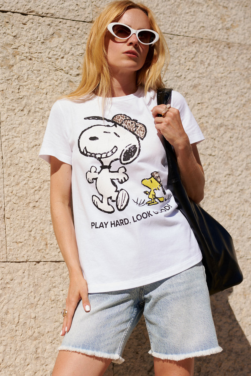 Eine Frau mit langen blonden Haaren, die eine weiße Sonnenbrille im Cat-Eye-Stil und ein weißes Snoopy-T-Shirt aus Baumwolle der „Frogbox by Princess Limited Edition“ sowie helle Jeansshorts trägt, steht mit einer schwarzen Umhängetasche vor einer beigen Wand.