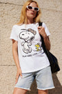 Eine Frau mit langen blonden Haaren, die eine weiße Sonnenbrille im Cat-Eye-Stil und ein weißes Snoopy-T-Shirt aus Baumwolle der „Frogbox by Princess Limited Edition“ sowie helle Jeansshorts trägt, steht mit einer schwarzen Umhängetasche vor einer beigen Wand.