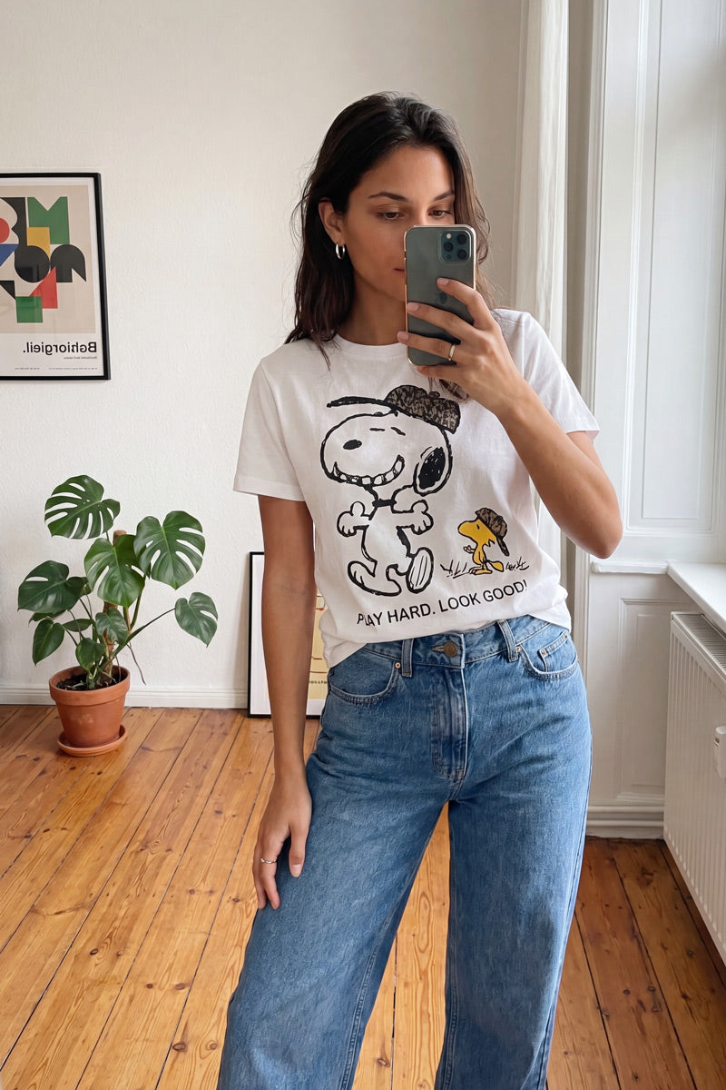 Eine Frau macht drinnen auf einem Holzboden ein Spiegel-Selfie. Sie trägt ein „Frogbox by Princess Limited Edition Snoopy“-T-Shirt, das sie in eine blaue Jeans mit hoher Taille gesteckt hat. Hinter ihr stehen eine Topfpflanze, ein abstraktes Bild und ein von Sonnenlicht durchflutetes hohes Fenster.