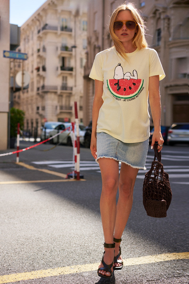 Eine Frau schlendert in einem „Frogbox by Princess Limited Edition Snoopy“-T-Shirt aus Baumwolle, kombiniert mit einem ausgefransten Jeansrock, durch eine Stadtstraße. Das Sonnenlicht spiegelt sich in ihrem blonden Haar, während sie eine geflochtene Tasche trägt und ihre schwarzen Sandalen mit Absatz auf dem Pflaster klappern.