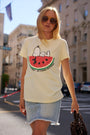 Eine Frau mit langen blonden Haaren und Sonnenbrille steht an einem sonnigen Tag draußen. Sie trägt ein hellgelbes Snoopy-T-Shirt aus der „Frogbox by Princess Limited Edition“-Kollektion, auf dem Snoopy auf einer Wassermelone zu sehen ist, dazu einen hellen Jeansrock und eine braune Stofftasche.