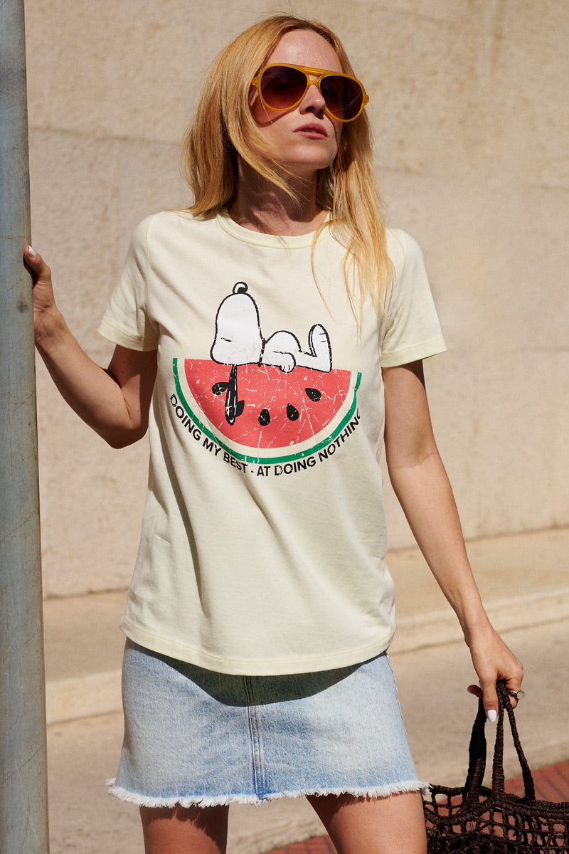 Eine Frau mit Sonnenbrille steht draußen mit einer braunen Netz-Tasche und einem Stock in der Hand; sie trägt ein hellgelbes „Frogbox by Princess Snoopy“-T-Shirt, auf dem Snoopy auf einer Wassermelonenscheibe zu sehen ist, dazu einen Jeansrock, vor einer strukturierten Steinmauer.